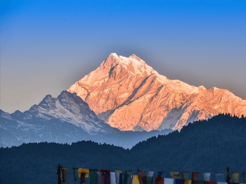 Kanchenjunga peak glowing orange at sunrise above dark forested ridges with prayer flags, Kanchenjunga Circuit Trek