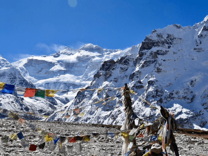 Colorful prayer flags on a rocky trail with snow-covered Kanchenjunga peaks under blue sky